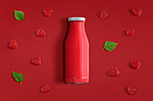 From above bottle of fresh juice of red color placed on dark red background with berries and green leaves