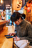 Asian lady in casual sweater using mobile phone at counter in traditional ramen bar