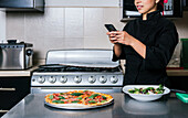 Crop unrecognizable female chef in uniform taking photo on smartphone of delicious cooked pizza and fresh vegetable salad in modern kitchen in restaurant