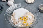 Mehl und Zucker in Glasschale mit Orangenschale für Blaubeermuffins auf Tisch mit Eiern während des Kochvorgangs