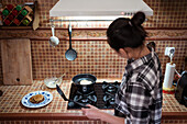 From above side view of focused young Asian female in casual clothes cooking yummy pancakes using spatula while standing in kitchen
