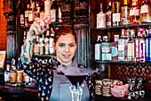 Zufriedene Barkeeperin in Schürze bei der Zubereitung eines Flavor-Blaster-Cocktails am Holztresen einer Bar