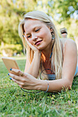 Smiling charming female lying on grass in park and listening to music in headphones in summer