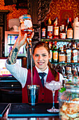 Smiling female barkeeper pouring alcohol in shaker while preparing refreshing cocktail at counter in bar and looking at camera