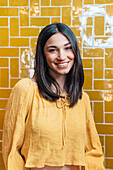 Young friendly female in yellow wear looking at camera with toothy smile against bright wall