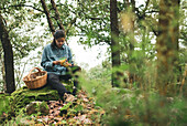 Serious female mycologist sitting on mossy boulder looking at Lactarius deliciosus mushroom in forest with basket