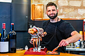 Bearded professional ethnic cutter preparing tasty jamon in restaurant with bottles of wine