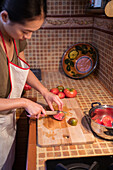 From above side view of ethnic female in apron cutting ripe tomatoes on chopping board while cooking lunch in kitchen at home