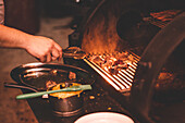 Crop faceless person with tongs grilling meat on rack in hot barbecue near sauce during cooking process in light cafe