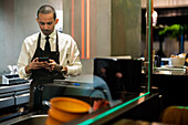 Seriöser Kellner in Uniform und Maske zum Schutz vor COVID 19 surft im Restaurant mit dem Handy durch das Glas