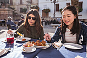 Content Asian woman with female friend eating fried nuggets while having lunch together at table in outdoor cafeteria on sunny day