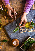 From above of crop lady cutting rosemary sprigs with scissors at table with fabric and rope near essential oil glass bottles and small chest