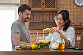 Young Asian lady with dark hair in casual clothes pouring olive oil from glass bottle into bowl with delicious healthy salad while preparing dinner near Hispanic husband in kitchen