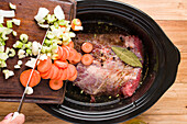 From above of crop anonymous person putting sliced carrot onion and celery from chopping board into slow cooker with beef with seasonings while preparing corned meat