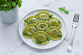Top view of tasty raviolis with basil decorated with pine nuts served on white round plate on table in light kitchen