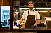 Junge männliche Köchin mit Schürze und Handschuhen, die ihr Gesicht mit einer Maske schützen, kocht in der Küche eines Restaurants und schaut in die Kamera