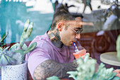 Through window side view of young cool male enjoying tasty healthy drink from disposable glass while looking down in daylight