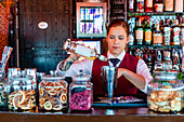 Female barkeeper pouring alcohol in shaker while preparing refreshing cocktail at counter in bar and looking away