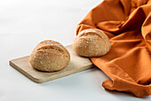 Fresh loaves of bread served on wooden chopping board near orange fabric on white background