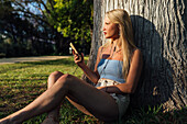 Unbeschwerte Frau, die mit ihrem Smartphone telefoniert und über Kopfhörer Musik hört, während sie bei Sonnenuntergang im Sommer unter einem Baum im Park sitzt