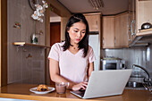 Focused Asian female with hot drink surfing netbook while sitting at counter with food during breakfast in modern kitchen at home