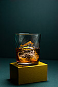 Close-up of a glass cup with cold whiskey and cubes of ice placed on a gold-colored rugged base and a turquoise blue corner background