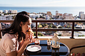 Seitenansicht einer asiatischen Frau, die an einem Tisch auf dem Balkon einer Wohnung sitzt und Gebäck isst, mit Blick auf eine Stadtlandschaft mit modernen Gebäuden