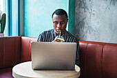 Attentive African American male entrepreneur with disposable glass of beverage sitting on sofa against table with netbook indoors