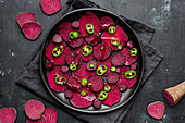 Top view composition of tasty beetroot slices arranged on baking pan with green jalapeno peppers and placed on black towel on kitchen table