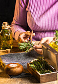Crop lady cutting rosemary sprigs with scissors at table with fabric and rope near essential oil glass bottles and small chest