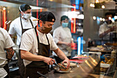 Through glass of focused male cook in uniform and protective mask adding ingredient to burger in kitchen with colleagues during coronavirus pandemic