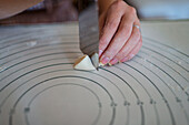 High angle of crop anonymous lady cutting raw dough with putty knife while preparing homemade dumplings sitting at table in kitchen