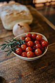 High angle of bowl with fresh cherry tomatoes near rosemary stems and whole onion on wooden table