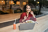 Blick durch eine Glasscheibe auf eine junge Bloggerin, die in einem modernen Restaurant an einem Tisch mit einer gesunden Mahlzeit sitzt und einen Laptop benutzt