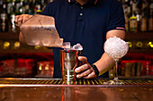 Hands of unrecognizable bartender putting ice cubes into the shaker while preparing a cocktail in the bar