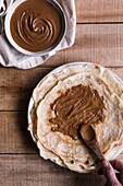 Von oben anonyme Person, die einen Löffel benutzt, um süße Nussbutter für leckere Crepes auf einem Holztisch zu verteilen
