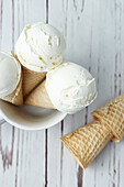 Top view of crispy waffle cones with scoops of yummy vanilla ice cream placed in cup on white wooden table in daytime