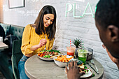 Cheerful female at table with appetizing meal and smoothies against crop anonymous ethnic partner in restaurant