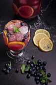 High angle of glass and jug with refreshing cold lemonade with fresh blueberries and lemon slices placed on dark table