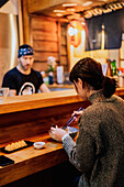 Side view of satisfied Asian woman in sweater smiling while taking spoon from worker sitting at wooden counter in ramen bar