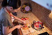 From above side view of ethnic female in apron cutting ripe tomatoes on chopping board while cooking lunch in kitchen at home