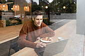 Blick durch das Fenster auf einen glücklichen jungen männlichen Studenten, der mit einem Laptop und einer Tasse Kaffee mit Croissant am Tisch sitzt und seine Zeit in einer modernen Cafeteria verbringt