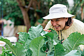 Delighted female gardener in hat smelling leaves of green cabbage growing in garden in village