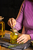 Crop anonymous lady in purple sweater showing essential oil glass bottles with herbs sprigs with green leaves near cloth on table
