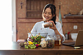 Positive young ethnic female with dark hair in casual clothes adding nuts into bowl of delicious salad and smiling while watching online recipe on laptop and cooking in kitchen