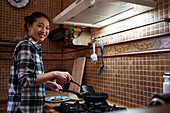 Side view of focused young happy Asian female in casual clothes cooking yummy pancakes using spatula while standing in kitchen looking at camera