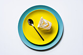 Draufsicht auf einen Joghurtbecher und einen silbernen Löffel auf gelben und blauen Keramiktellern auf einem weißen Tisch in der Küche
