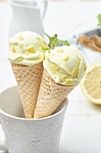 Pair of yummy homemade lemon ice cream in crispy waffle cones decorated with zest and mint leaf placed in cup on table in kitchen