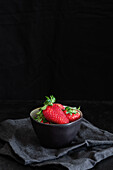 Tasty fresh strawberries with sepals in round shaped bowl on crumpled textile on black background