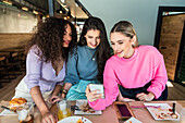 Smiling young female friends wearing casual clothes browsing mobile phones while gathering for lunch in restaurant